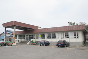 茶色の屋根に外壁がベージュ色をした外観で建物前が駐車スペースとなっているつがる市地域包括支援センターの外観写真