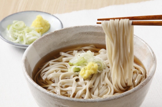 ネギとすりおろしたショウガが添えられたお椀に入ったうどんを箸で持ち上げている写真