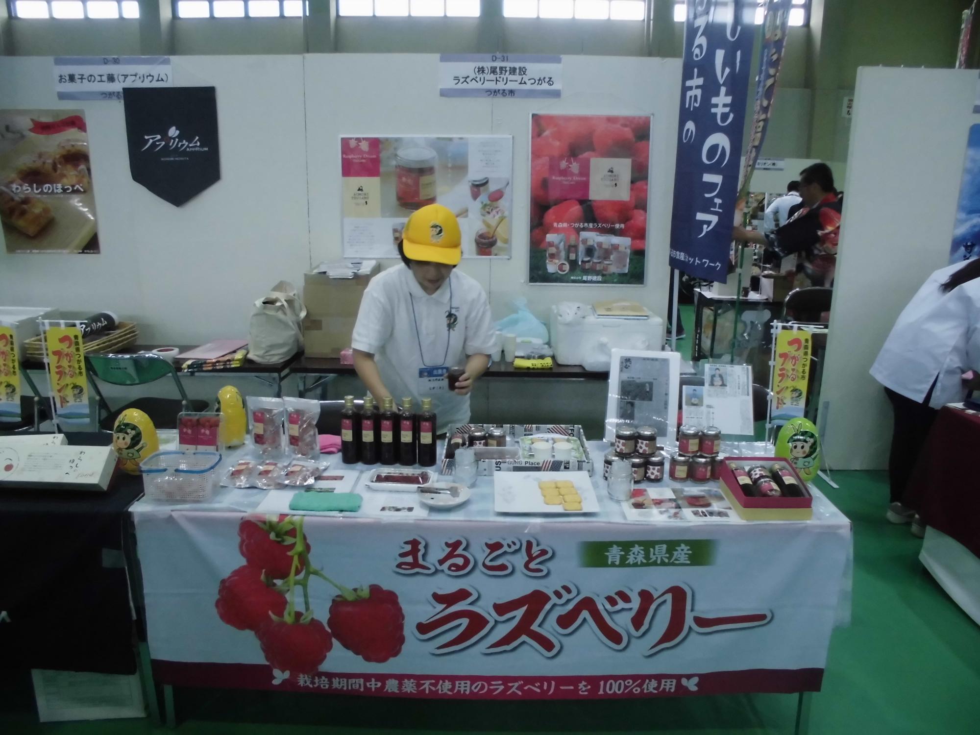 「青森県産まるごとラズベリー」とラズベリーの写真が印刷された暖簾がブースのテーブルに掲示され、いろいろな種類の商品を職員の女性が机の上に並べている商談会の様子の写真