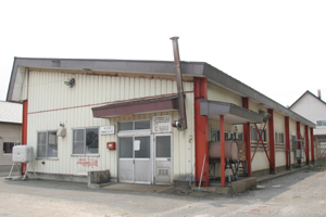 クリーム色の壁にアクセントで赤が使われ、入り口横に煙突があるつがる市稲垣農産物加工センターの外観を入り口を正面にして写した写真