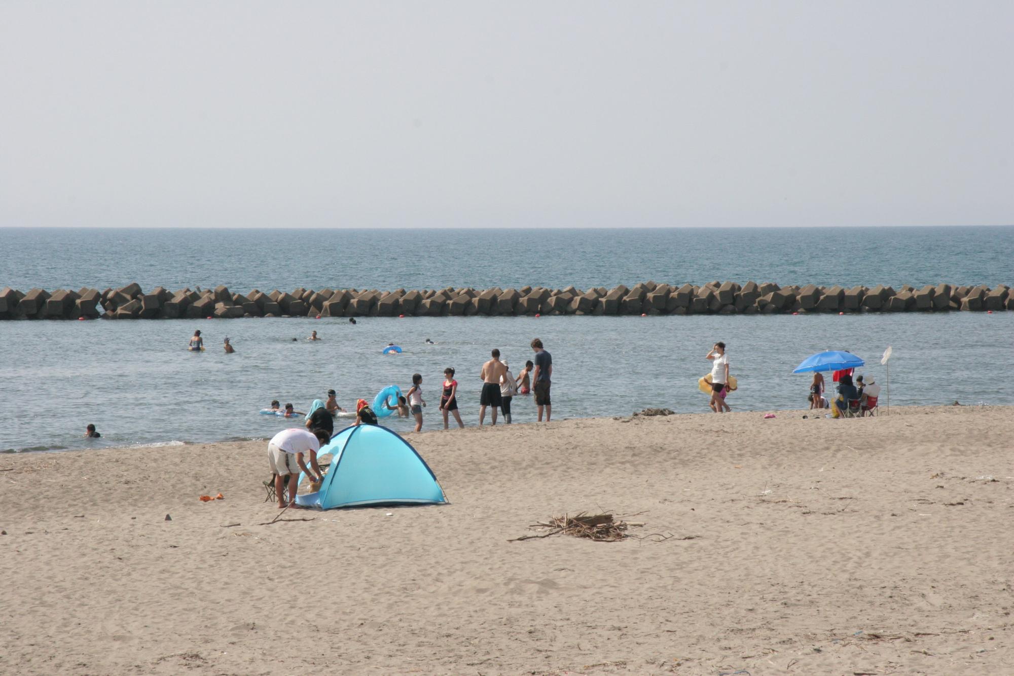 砂浜に水色のテントを広げ、奥には家族連れが楽しんでいる観光客で賑わう出来島海水浴場の写真