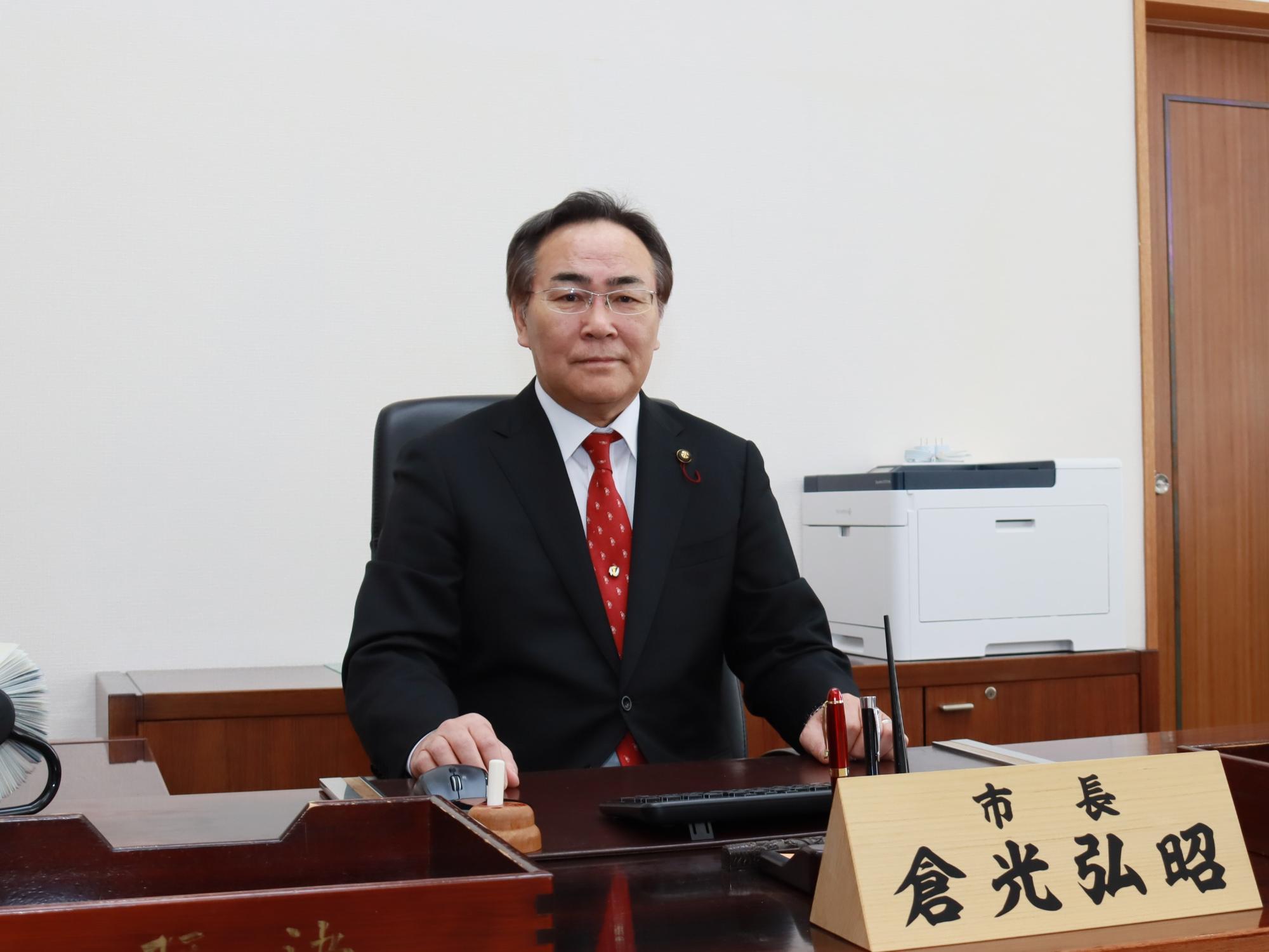 市長デスクに置かれた卓上名札の前で椅子に座っている倉光弘昭市長の写真