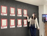 壁に展示されている8枚の広告ポスターの横に女性が立ち、1枚のポスターを指して紹介している様子の写真
