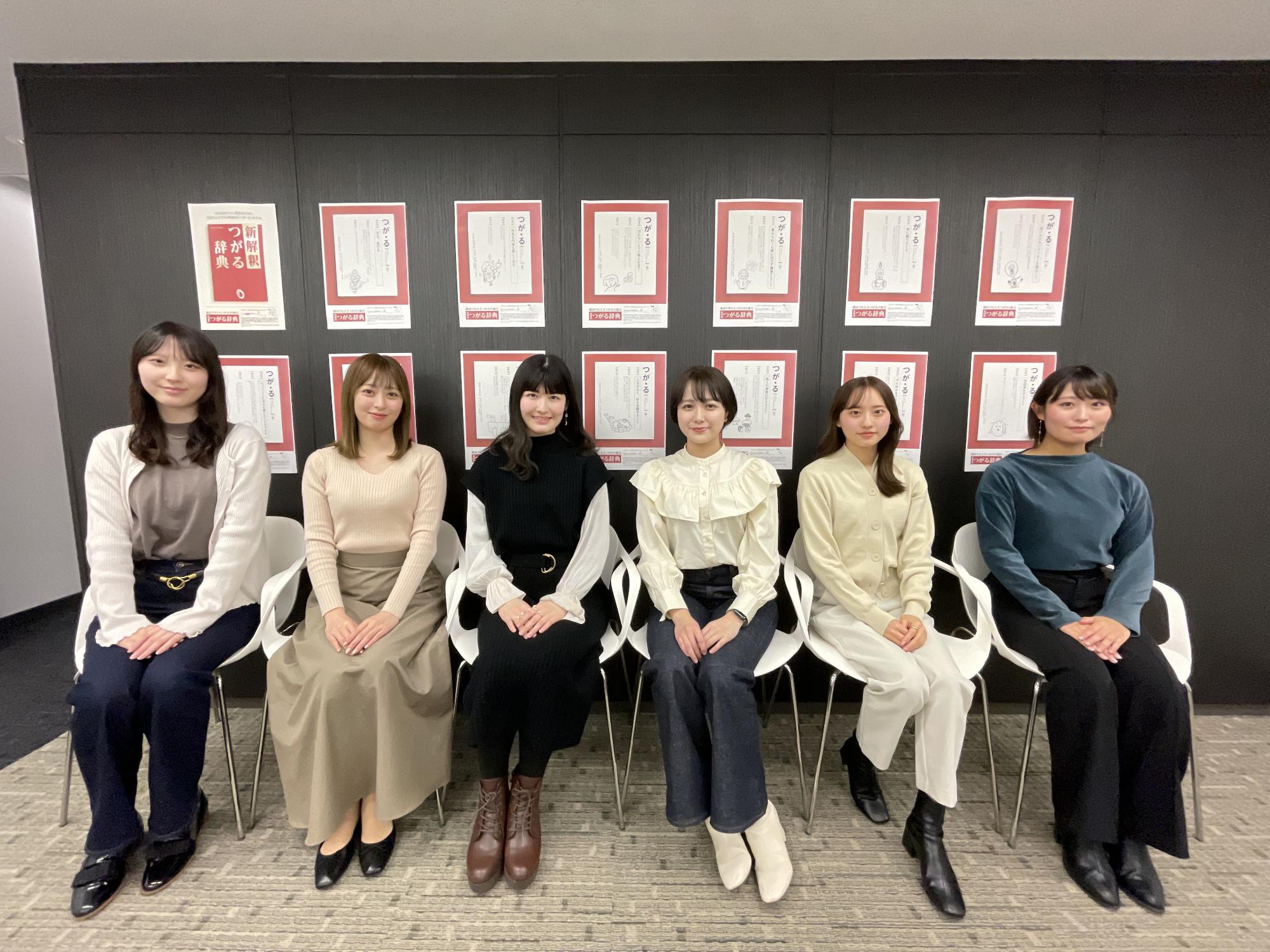 壁に展示されている広告ポスターをバックに6名の女性たちが椅子に座り記念撮影をしている写真