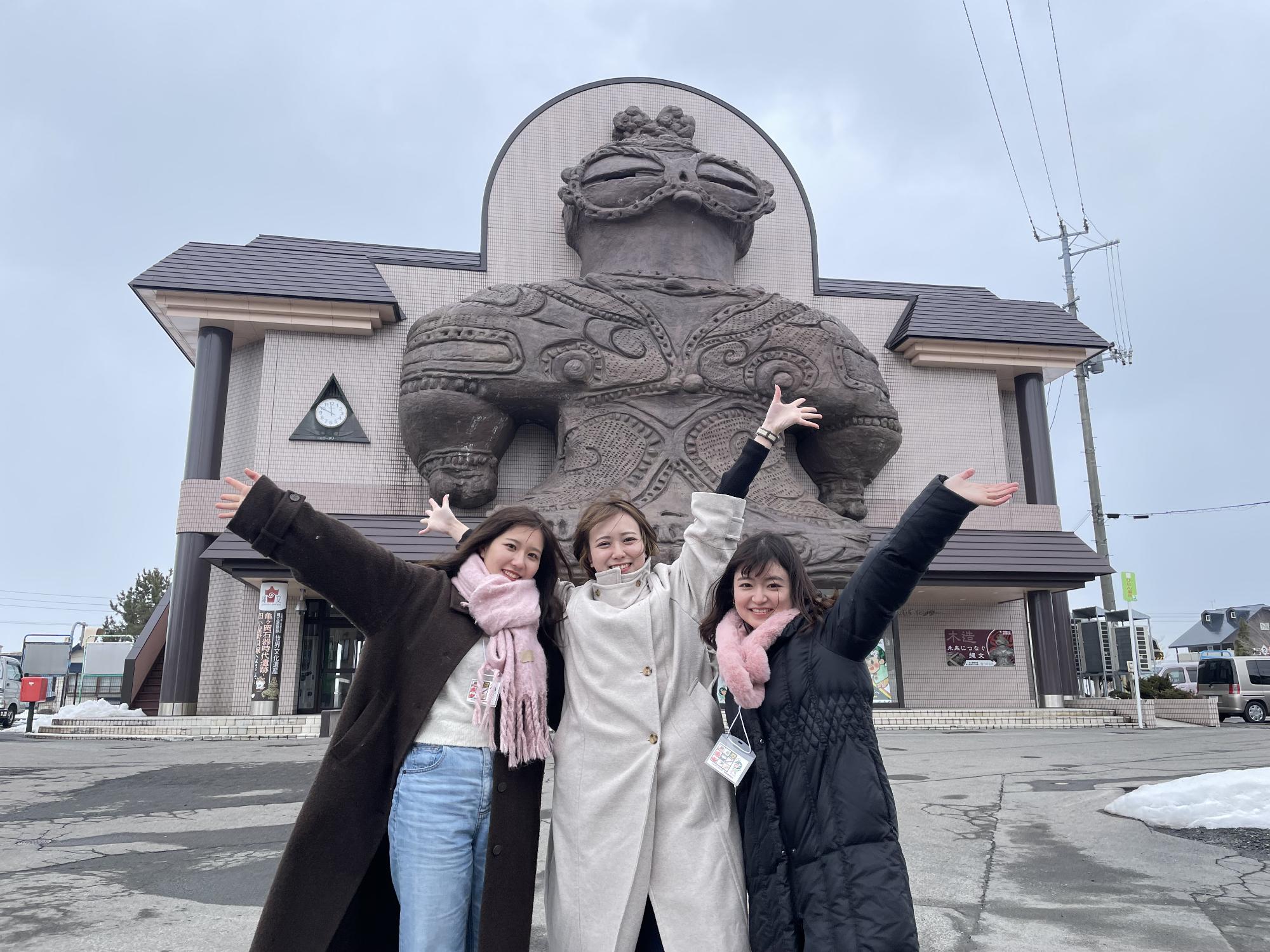 大きな遮光器土偶のモニュメントの前で学生3人が手を上に広げてポーズをとっている写真