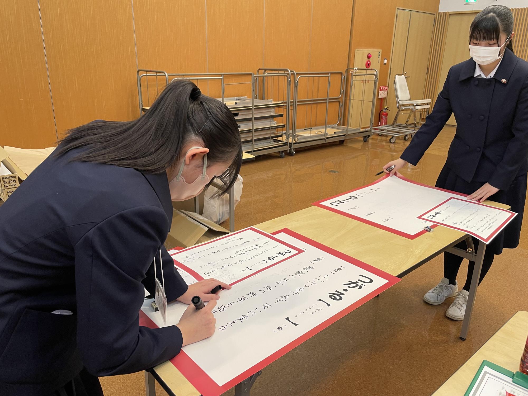 長机の上で、赤い紙の台紙の上に貼られた紙に文字を書いている女子学生の写真