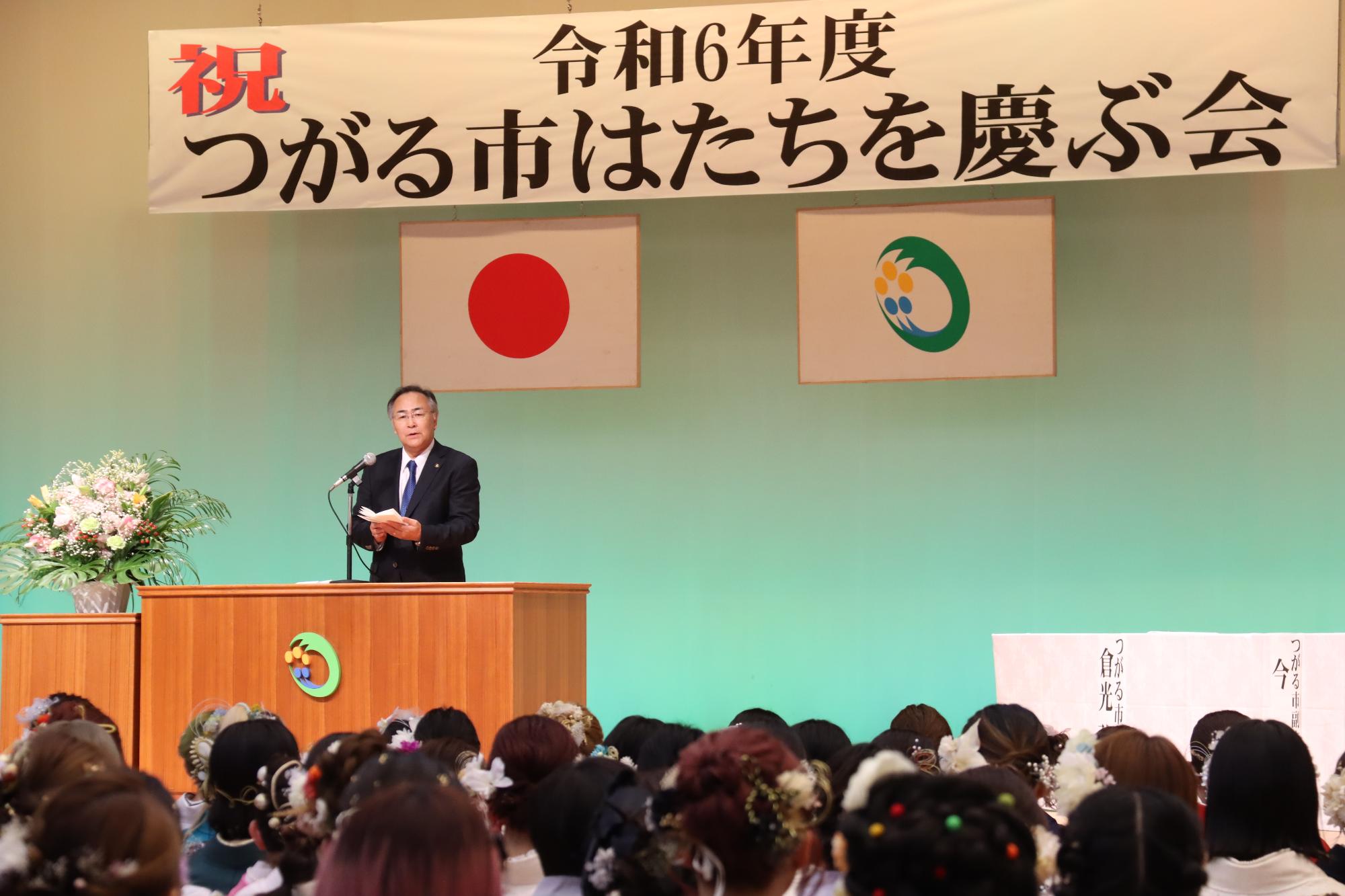 壇上に吊るされた「つがる市はたちを慶ぶ会」と書かれた横断幕を背景に市長が祝辞を述べているところを参加者が視聴している写真