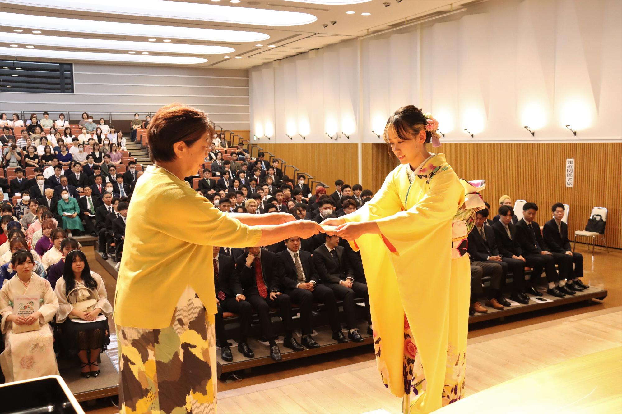 参加者が見守る中、壇上で関係者より記念品が贈呈され振り袖姿の女性が受け取っている様子の写真