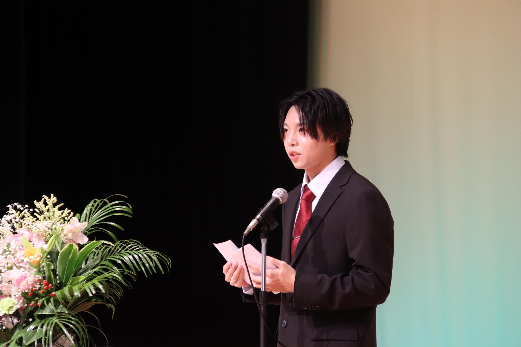 壇上に置かれたマイクの前でスーツ姿の男性が奉書紙を両手に持ち話をしている写真