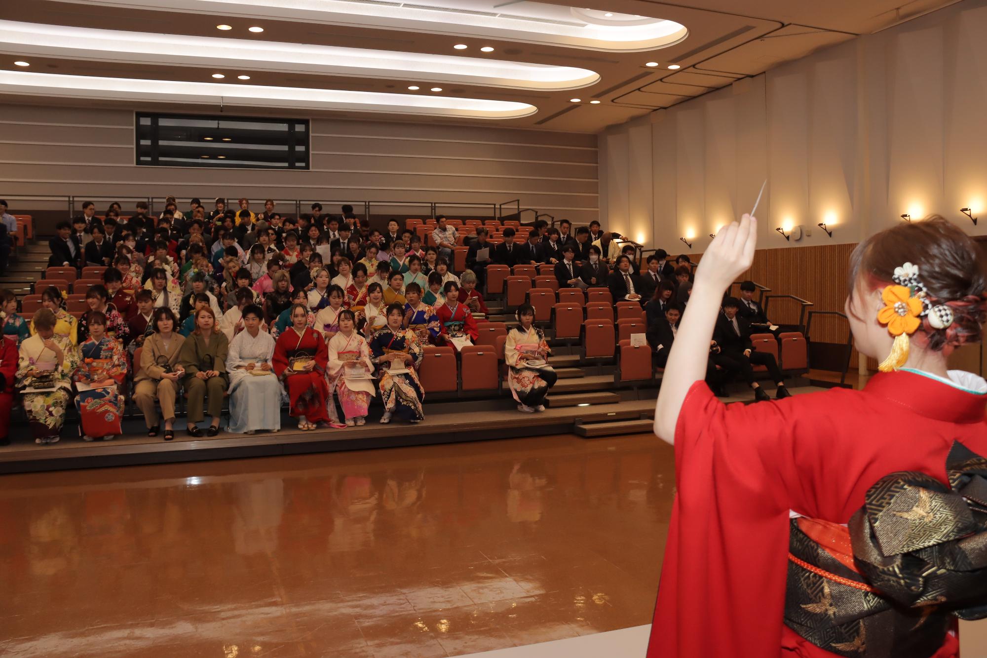 赤い振袖を着た女性が観覧席に座っている参加者に向けて手を挙げ紙を見せている抽選会の写真