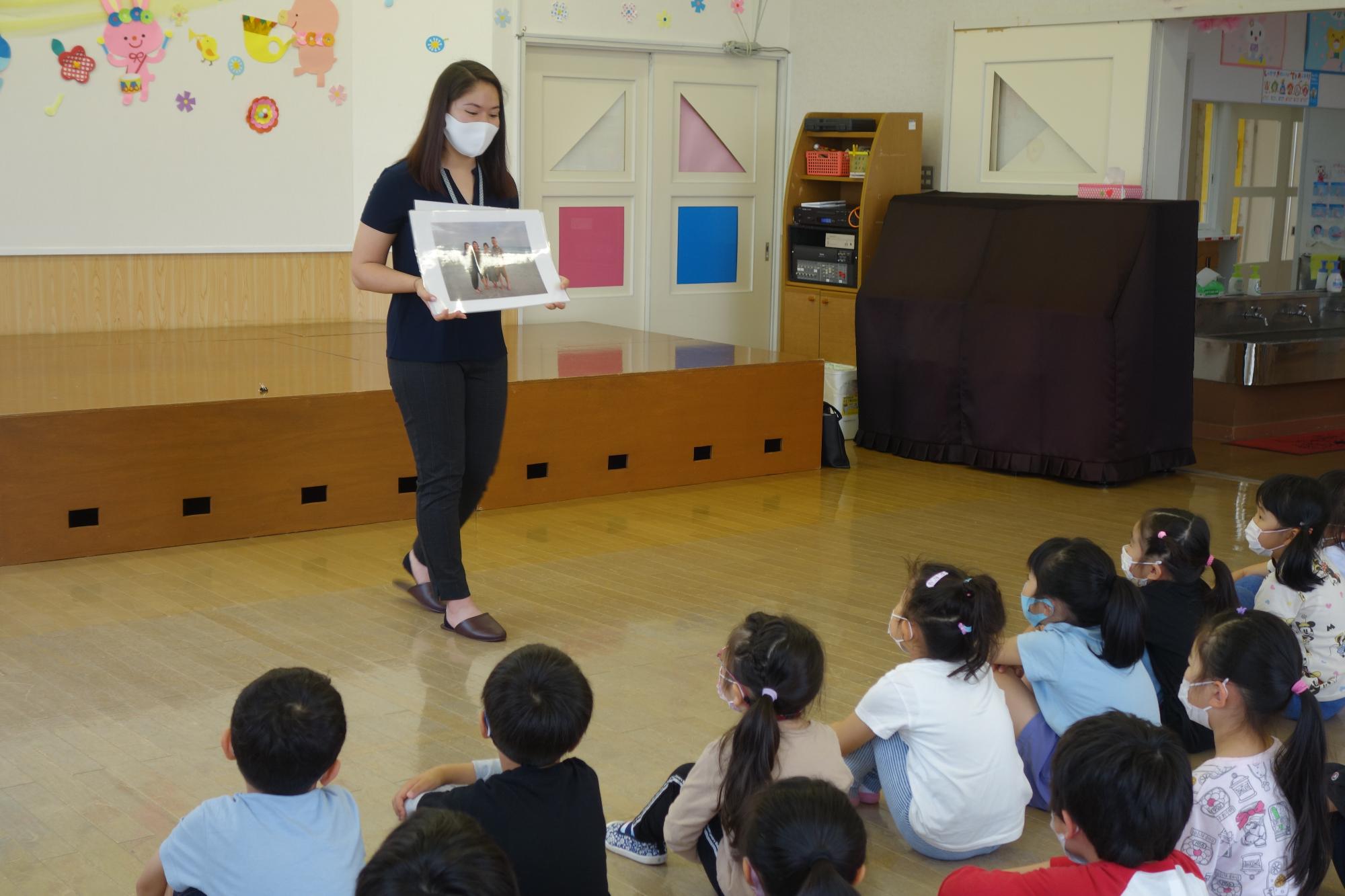 小さなステージが設置された室内の広場の床に園児たちが体育座りし、前方に立つ国際交流員のオリビアさんが大きく引き伸ばした家族の写真を持って園児に見せている様子の写真