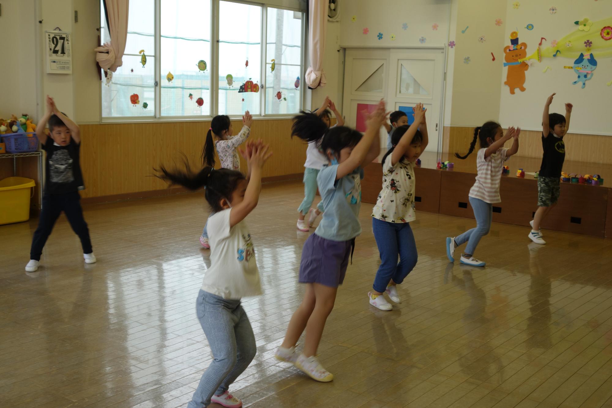 教室内を2列に並び間隔をあけて広がっている園児たちが、両手を上に挙げて踊っている様子の写真
