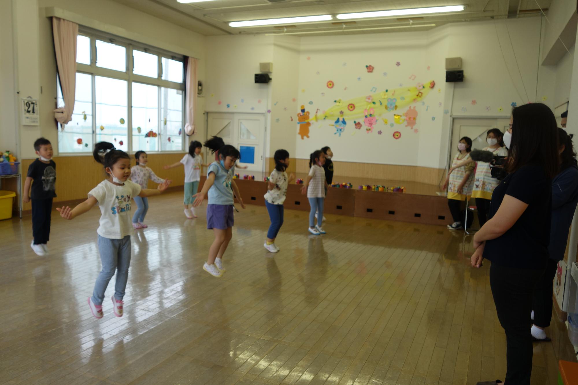 教室内を2列に並び間隔をあけて広がって踊っている園児たちを、先生たちやオリビアさんが見ている写真
