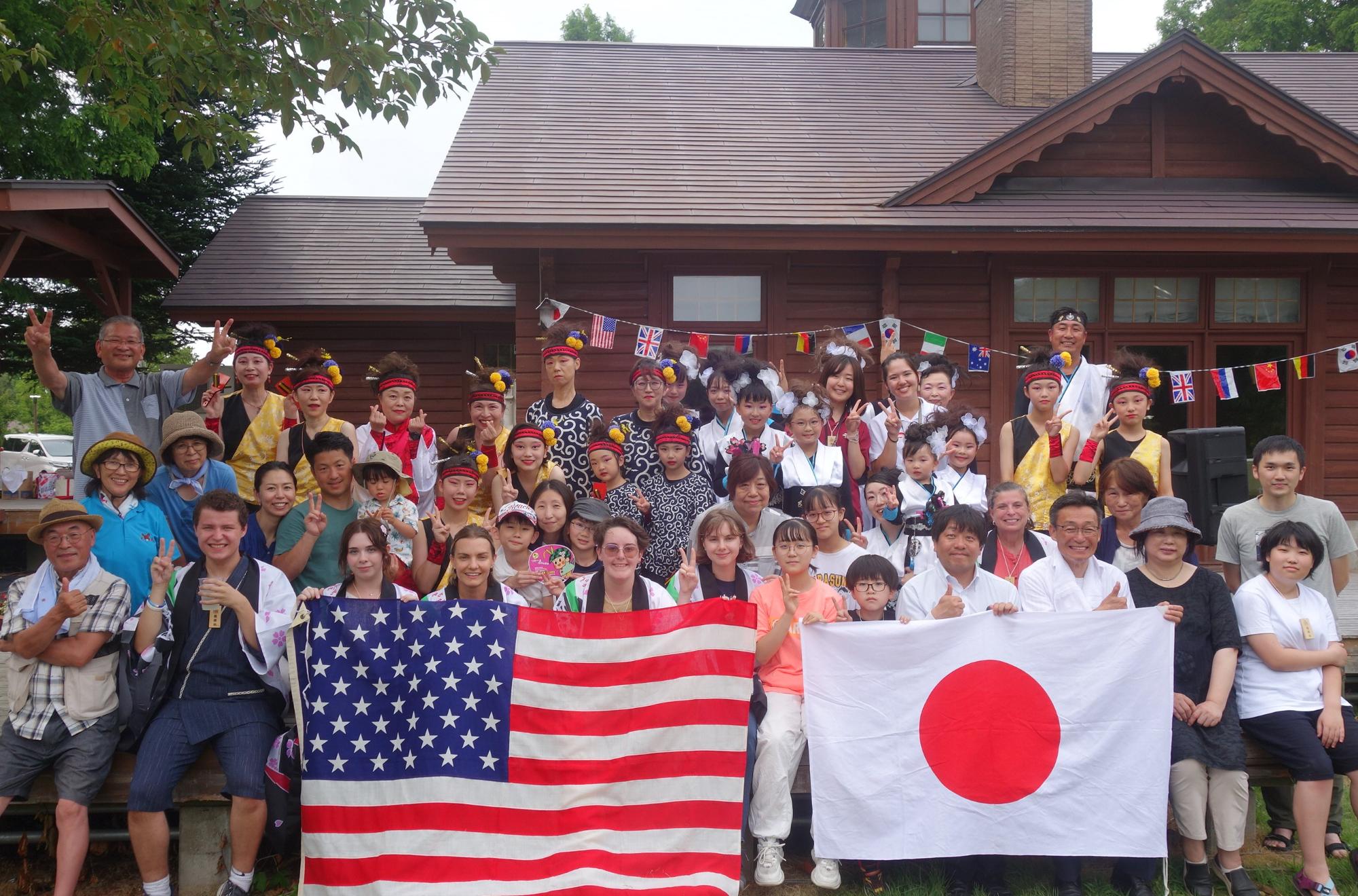 ログハウスの前でホストファミリーや参加者そして訪問団が整列し、前列のメンバーがアメリカと日本の国旗をそれぞれ両手に持ちそれぞれポーズを取りながら記念撮影をしているさよならパーティの様子を撮影した写真