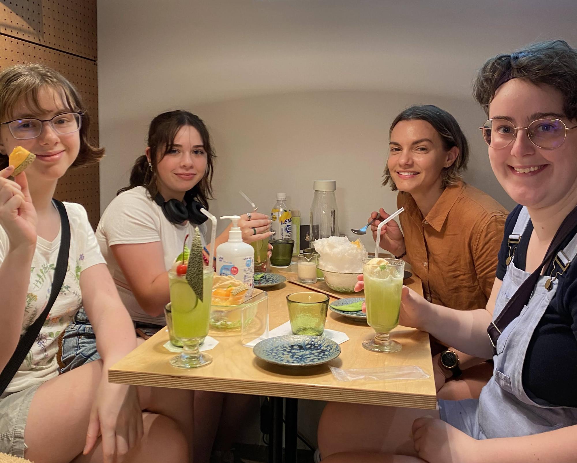 かき氷やメロンジュースなどの食べ物が乗ったテーブルの前で笑みを浮かべる訪問団4名の女性たちの写真