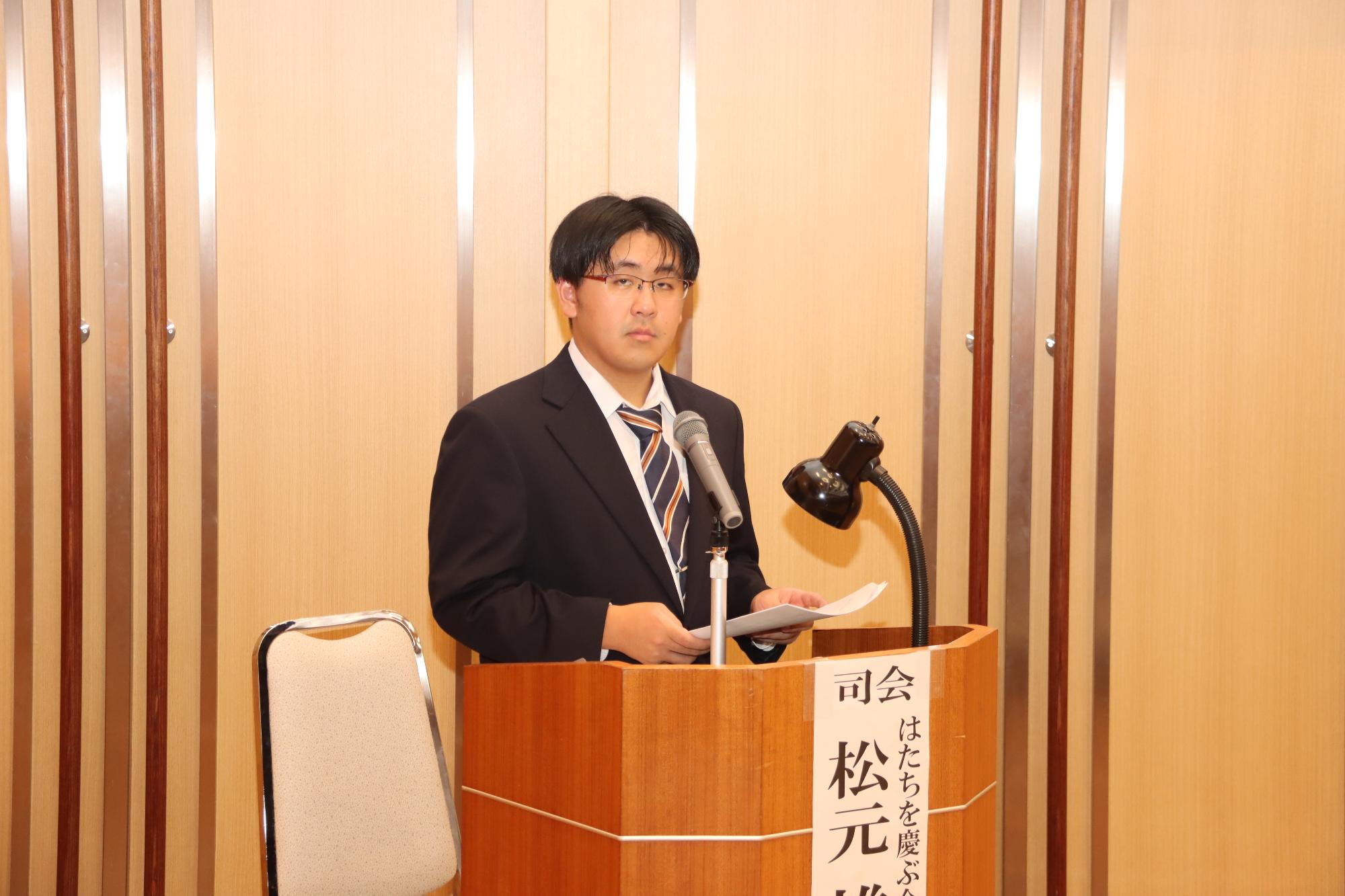 スーツを着た男性がマイクの前で式典の司会進行をしている様子