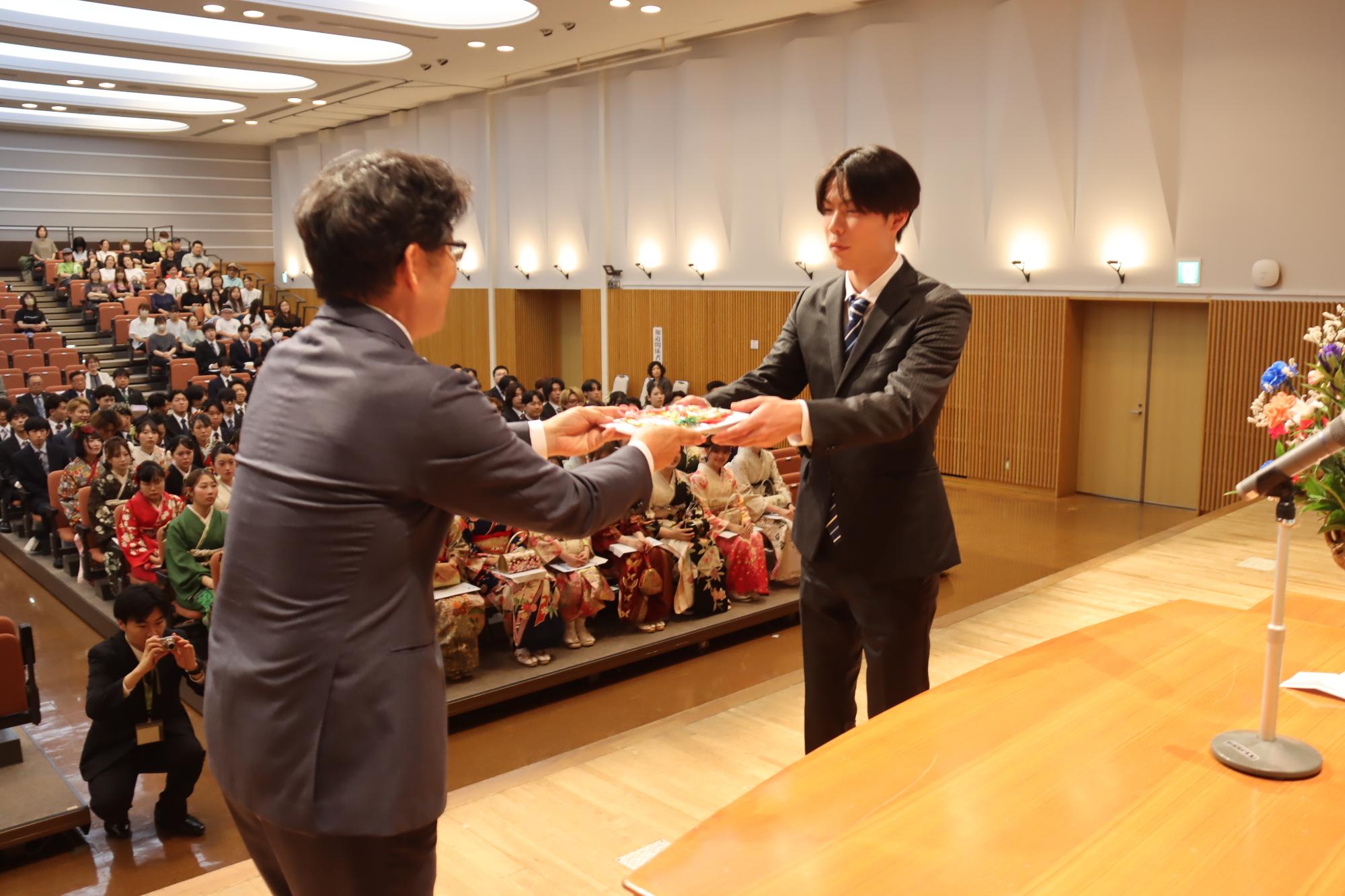 参加者が見守る中、壇上で関係者より記念品が贈呈されスーツ姿の男性が受け取っている様子の写真
