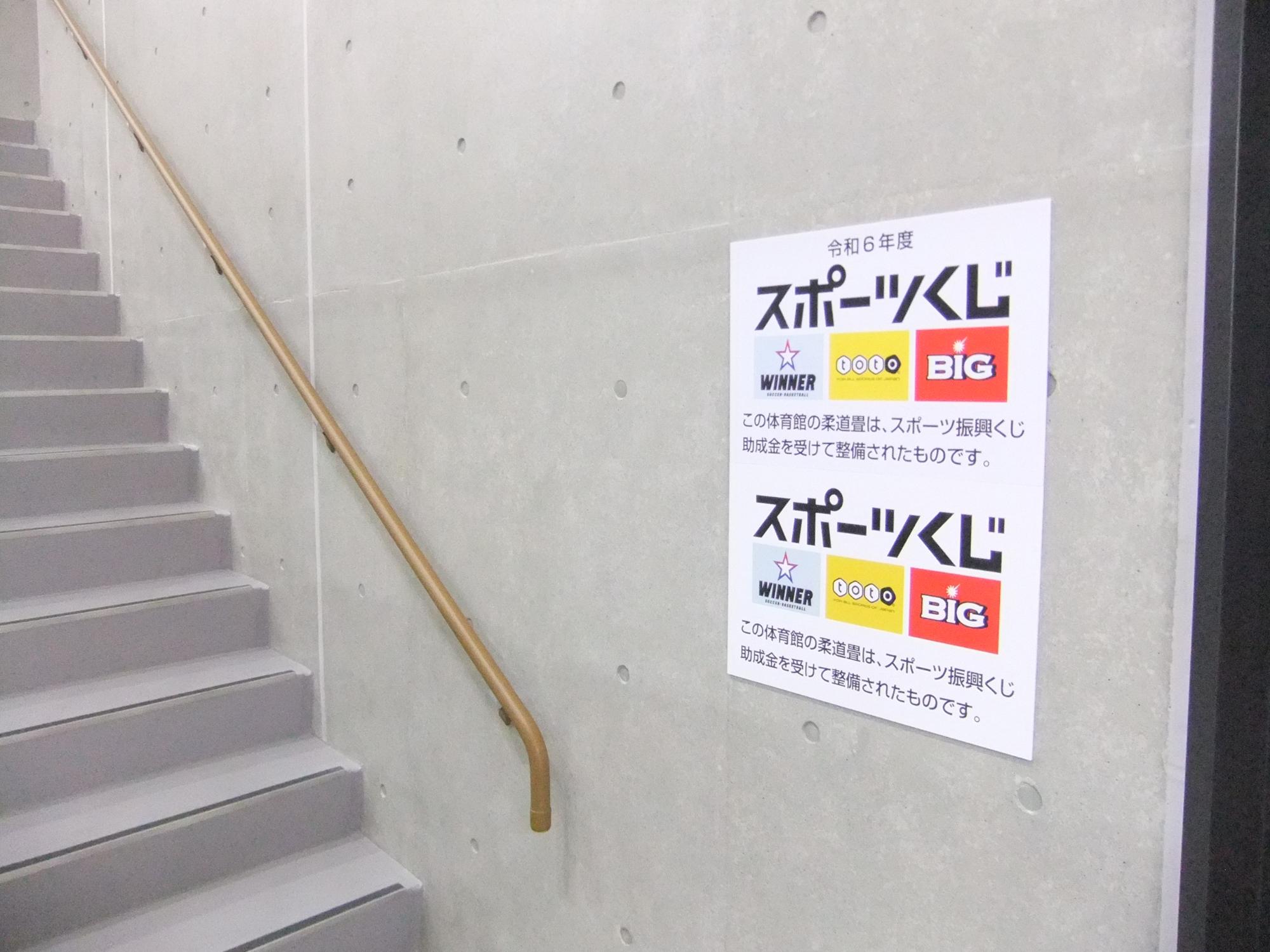 スポーツくじのロゴと共に「この体育館の柔道畳は、スポーツ振興くじ助成金を受けて整備されたものです。」と書かれたポスターが壁に掲示されている写真