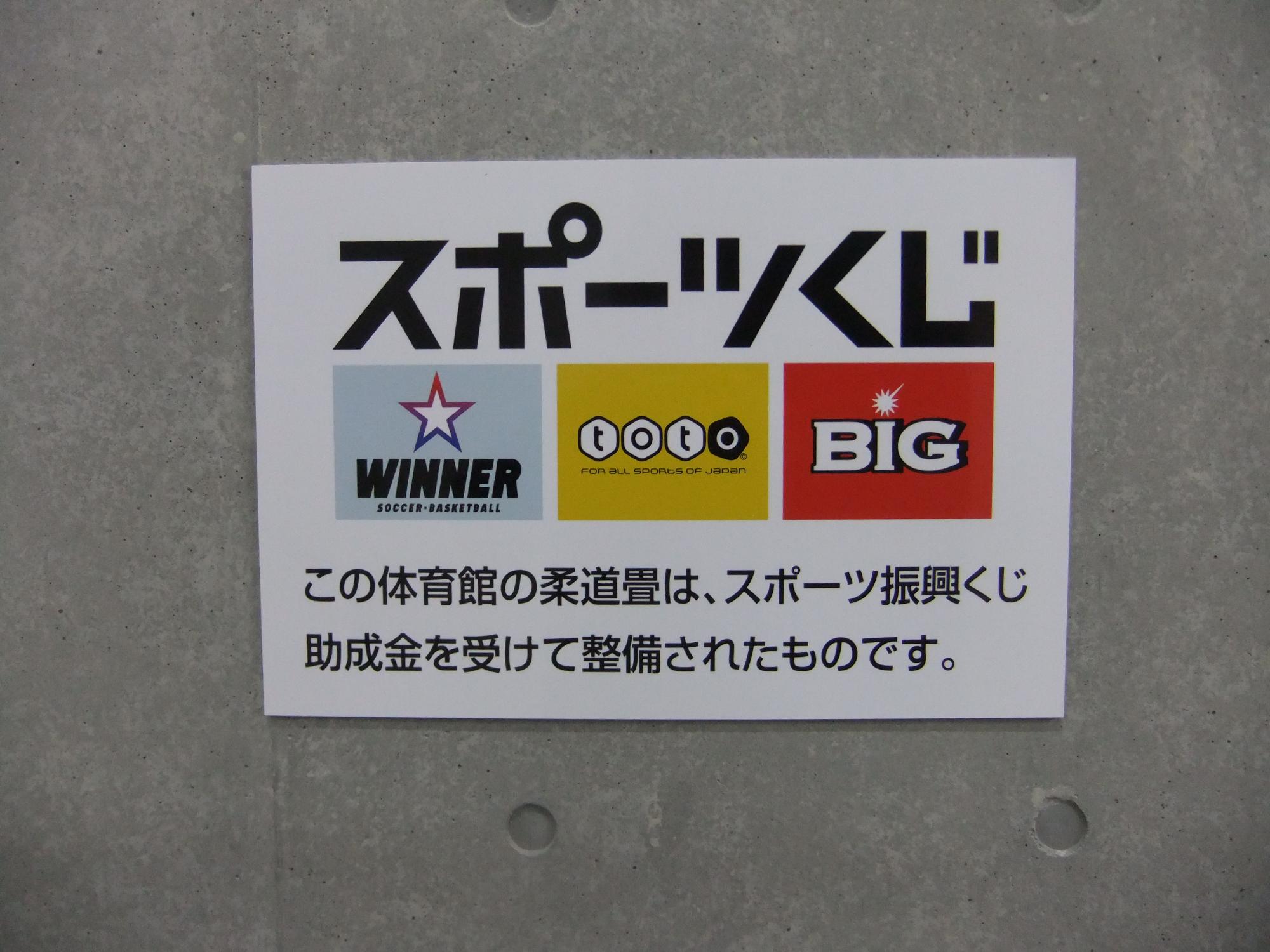 スポーツくじのロゴと共に「この体育館の柔道畳は、スポーツ振興くじ助成金を受けて整備されたものです。」と書かれたポスターが壁に掲示されている写真